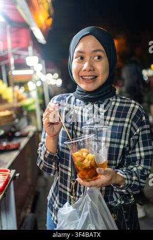 Indonésienne asiatique savourant une nourriture de rue locale sur un marché de nuit animé à Yogyakarta. Concept de tourisme culinaire, culture alimentaire et vie nocturne. Banque D'Images