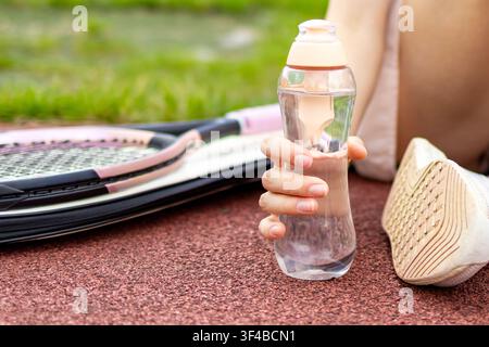 Close-up d'une main tenant une bouteille d'eau réutilisable à côté d'une raquette de tennis après un entraînement en extérieur sur un terrain de sport. Concept d'hydratation, de récupération, de durabilité Banque D'Images