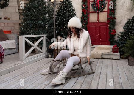 Scène de vacances d'hiver mettant en scène une femme en tenue blanche duveteuse assise sur un traîneau en bois vintage sur un porche décoré Banque D'Images