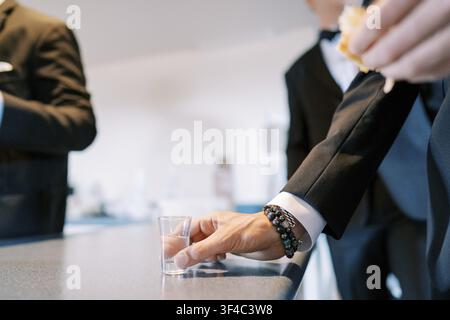 Meilleur homme tenant un verre à shot près du comptoir du bar, manche de smoking et montre-bracelet dans le cadre, main stable au-dessus du verre, invités flous en arrière-plan Banque D'Images