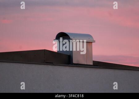 Cheminée de ventilation moderne sur le toit, silhouettée contre un ciel rose doux au coucher du soleil. Détail architectural minimal avec espace pour du texte. Banque D'Images