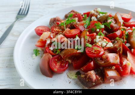 Salade de tomates de formes et de couleurs différentes, ail, piment, persil et huile d'olive avec une fourchette sur un fond en bois blanc. De la ferme à la table Banque D'Images