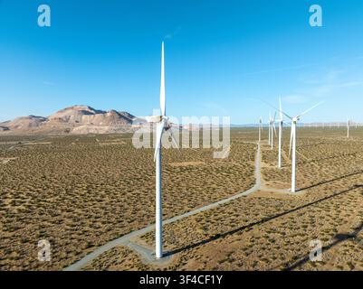 Vue aérienne de turbines éoliennes dans une ferme d'énergie propre du désert de Mojave avec un terrain aride, une route de terre et des montagnes sous un ciel bleu clair à Mojav Banque D'Images