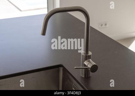 Un robinet en métal brossé contemporain avec un bec courbé et une poignée à levier unique est installé sur un plan de travail de cuisine sombre et lisse à côté d'un évier Banque D'Images