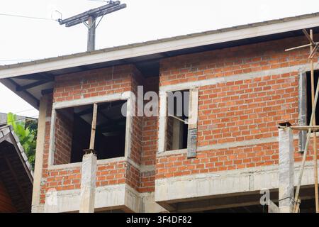 Une maison en briques inachevée en construction avec un mur de briques rouges apparent et des ouvertures de fenêtres montrant des échafaudages et des poutres en béton, lumière neutre du jour Banque D'Images