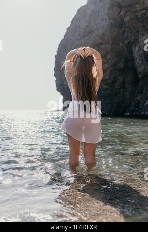 Femme de l'océan, jeune femme sur falaise, debout dans l'eau scintillante au pied de falaises de pierre escarpées pendant des vacances tranquilles sur la plage en été Banque D'Images