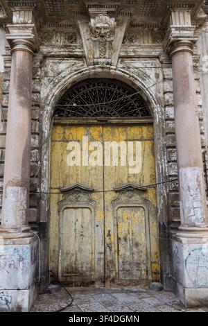 Une porte d'entrée en bois jaune vintage ornée, encadrée de colonnes en pierre et d'une porte en arc comportant une clé de voûte en forme de masque grotesque sculpté et un fanlight en fer finement ouvragé Banque D'Images