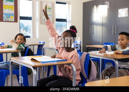 Enfants afro-américains assis en classe, une fille en pull rose levant la main avec un crayon Banque D'Images