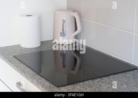 Comptoir de cuisine moderne avec une plaque à induction en verre noir, une bouilloire électrique blanche et un rouleau d’essuie-tout sur une surface en granit Banque D'Images