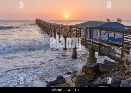 Un coucher de soleil doré éclatant encadre la jetée en bois historique de Swakopmund, en Namibie, alors que les puissantes vagues de l'océan Atlantique s'écrasent contre ses piliers. Banque D'Images