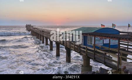Un coucher de soleil doré éclatant encadre la jetée en bois historique de Swakopmund, en Namibie, alors que les puissantes vagues de l'océan Atlantique s'écrasent contre ses piliers. Banque D'Images