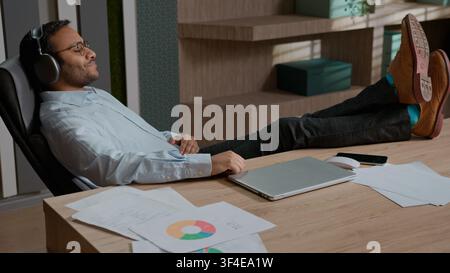 Homme d'affaires indien arabe détendu écoutant de la musique avec des écouteurs sans fil au bureau, homme d'affaires profitant d'une pause après le travail, calme Banque D'Images