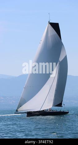 Vue d'un voilier de course élégant avec une grande voile blanche glissant sur les eaux bleues du lac Léman sous un ciel dégagé à Morges, Vaud, Suisse. Banque D'Images