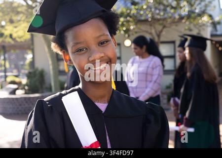 Jeune diplômé afro-américain portant une robe souriant tenant un diplôme avec un ruban rouge sur le campus Banque D'Images