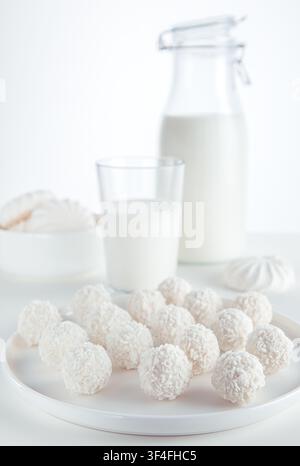 Bonbons truffes au coco blancs dans une assiette avec un verre et une bouteille de lait ainsi que des guimauves dans un bol sur un fond clair. Banque D'Images