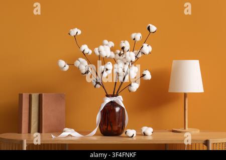 Branches de coton dans un vase en verre marron avec un ruban blanc sur une table en bois avec des livres et une lampe, le fond présente un mur orange uni, provid Banque D'Images