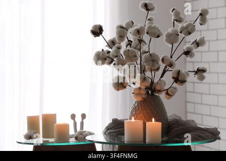 Branches de coton dans un vase décoratif avec des bougies allumées sur une table en verre, créant une décoration intérieure chaleureuse et naturelle Banque D'Images