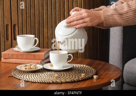 Main de femme versant du thé chaud d'une théière en céramique blanche dans une tasse sur une table en bois avec des morceaux de sucre, Des morceaux de sucre brun sont disposés sur un petit blanc Banque D'Images