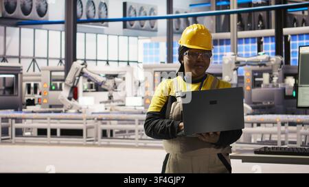 Spécialiste souriant de l'usine intelligente testant des systèmes automatisés pour garantir la fiabilité. Ingénieur heureux dans une usine industrielle 4.0 identifiant les goulots d'étranglement et optimisant les engrenages sur la base des données, caméra A Banque D'Images