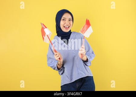 Jeune femme indonésienne heureuse portant un hijab tenant des drapeaux devant un fond mural jaune avec une mise au point nette pour le marketing de la fête de l'indépendance nationale Banque D'Images