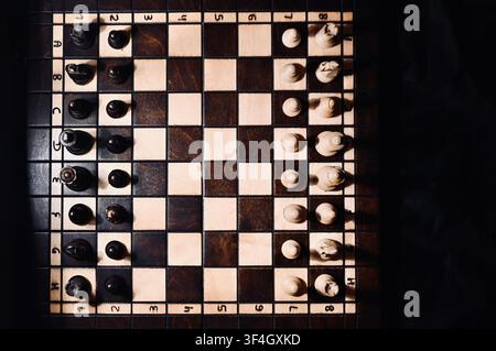 Vue en plongée d'un échiquier en bois avec toutes les pièces disposées en position de départ. Le contraste dramatique entre les nuances claires et sombres avec des ombres subtiles symbolise la planification stratégique, la concurrence commerciale, la vision tactique, la prise de décision et le défi intellectuel. Banque D'Images