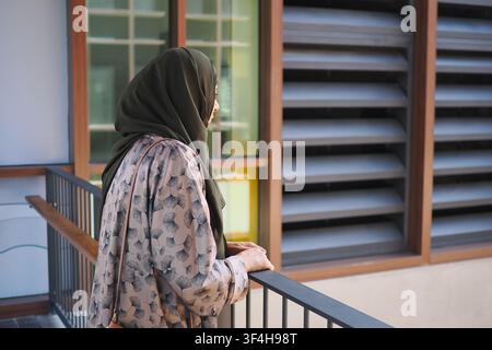 Femme musulmane contemplative en hijab debout sur un balcon surplombant une architecture urbaine moderne Banque D'Images