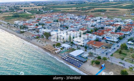 Paralia Dionisiou, Grèce, 16 juillet 2026. Des maisons basses en bord de mer, des parasols de plage et une eau calme bordent une plage de sable, vus du dessus à côté d’un f cultivé Banque D'Images