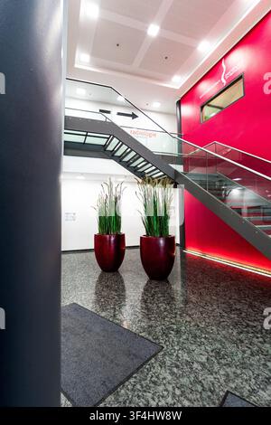 Hall d'entrée moderne au bureau avec mur d'accent rouge et escalier en verre, intérieur d'entreprise contemporain avec sol en granit, colonnes cylindriques et grande plante Banque D'Images