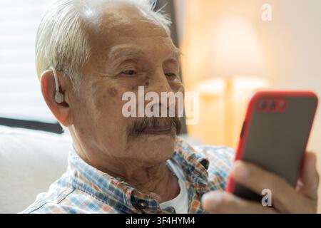 Grossissement vue latérale d'un homme âgé avec appareil auditif utilisant le téléphone à la maison. Mise au point sélective. Concept d'oreille avec appareil auditif BTE. Banque D'Images