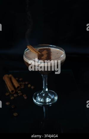 Cocktail de café crémeux garni de poudre de cacao et d'une bâtonnette de cannelle, servi dans un verre à cocktail élégant sur un fond sombre. Banque D'Images