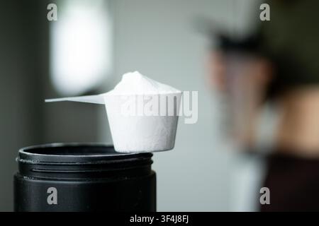 Cuillère à poudre protéinée blanche reposant sur un récipient noir avec un fond flou d'une personne tenant une bouteille shaker dans un environnement de fitness Banque D'Images