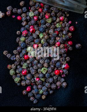 Poivrons mélangés colorés dispersés sur un fond sombre avec une cuillère en bois rustique remplie de poivrons entiers. Banque D'Images