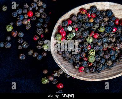 Poivrons mélangés colorés dispersés sur un fond sombre avec une cuillère en bois rustique remplie de poivrons entiers. Banque D'Images