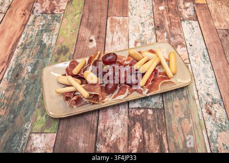 Plateau de jambon espagnol séché accompagné de raisins rouges et de baguettes, élégamment présenté sur des planches de bois usées. Banque D'Images