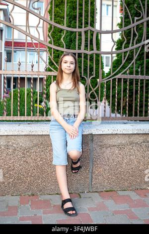 Adolescente assise sur un rebord de pierre près d'une clôture décorative en tenue d'été décontractée Banque D'Images
