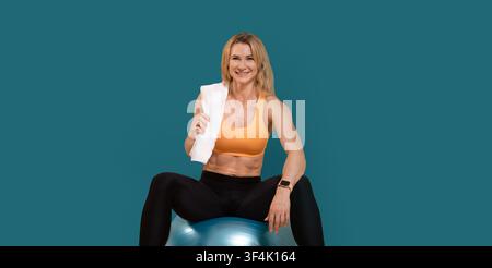 Détendez-vous après un entraînement de remise en forme à la maison. Femme d'âge moyen, musclée et souriante, vêtements de sport avec serviette sur l'épaule Banque D'Images