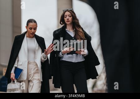 Deux femmes d'affaires marchant et discutant du travail tout en tenant une tablette et un carnet en plein air Banque D'Images