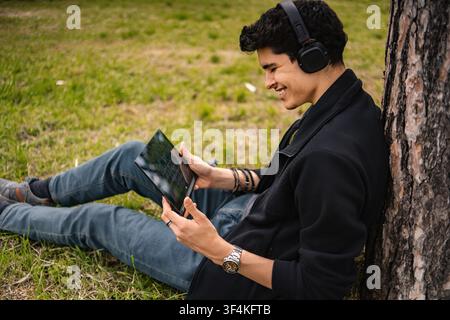 Jeune homme portant des écouteurs se détendant dans le parc en utilisant une tablette numérique Banque D'Images