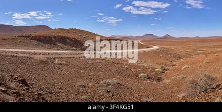 Une vue panoramique haute résolution capture une route de gravier clair serpentant à travers le terrain désertique rocheux et rouge de la Namibie vers des montagnes lointaines. Banque D'Images