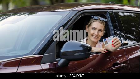 Femme souriante assise dans la voiture et montrant son nouveau permis de conduire par la fenêtre à la caméra Banque D'Images
