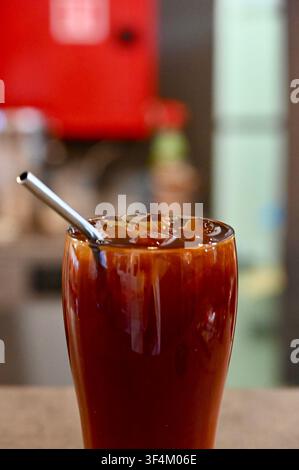 Un verre rafraîchissant de café américain glacé noir rempli de glaçons et d’une paille en métal réutilisable, posé sur un comptoir en bois dans un café moderne. Parfait pour les menus de cafés, les concepts de boissons estivales et les visuels lifestyle. Banque D'Images