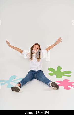 Un enfant joyeux est assis par terre, les bras grands ouverts, entouré de formes vives, embrassant l'amusement. Banque D'Images