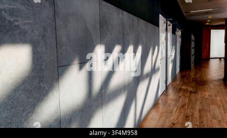 La lumière du soleil projette des ombres géométriques dramatiques le long d'un mur carrelé gris dans un couloir moderne vide, photographié depuis un angle diagonal bas. La scène Banque D'Images