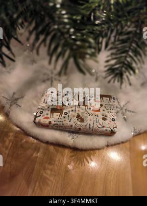 Un cadeau de Noël magnifiquement emballé avec des motifs d'animaux festifs, placé sous un sapin de Noël sur une jupe en fausse fourrure blanche. Banque D'Images