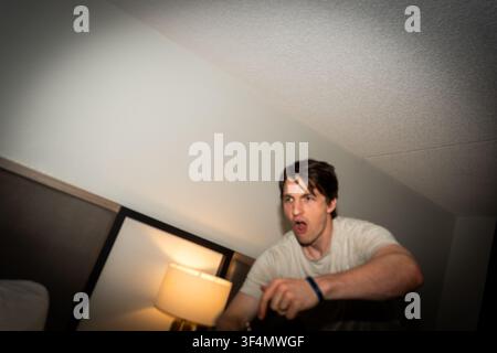 homme dans une chambre avec une expression surprise, gros plan, angle légèrement bas, plafond texturé, lit en bois. Banque D'Images
