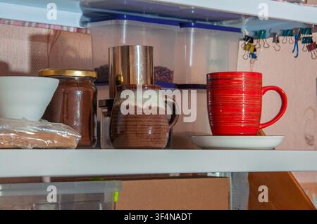 Une vue rapprochée d'une étagère de cuisine présentant divers articles, dont un pot de marc de café, deux tasses et des contenants de stockage alimentaire en plastique transparent Banque D'Images