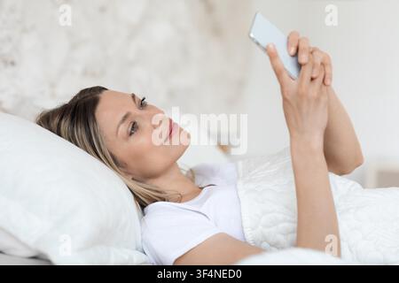 Jeune femme allongée dans son lit utilisant son smartphone à la maison, mode de vie détendu et confortable Banque D'Images