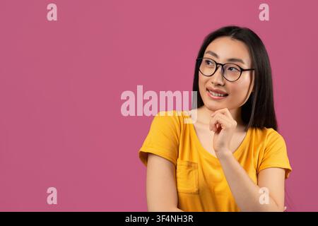 Femme asiatique pensive portant des lunettes, souriant et réfléchissant devant un fond rose, concept de style de vie Banque D'Images