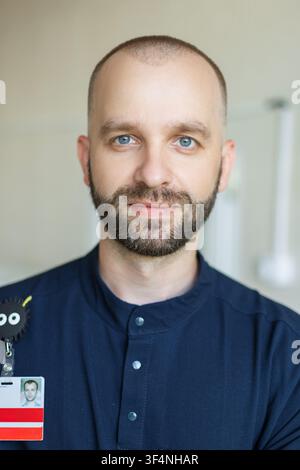 Portrait d'un médecin ou d'un infirmier masculin amical. Homme caucasien aux yeux bleus, barbu et à la tête chauve, portant une blouse médicale bleue et un badge d'identification. regardant Banque D'Images