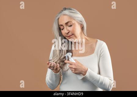 Femme âgée caucasienne utilisant du spray capillaire sur de longs cheveux gris, cadre intérieur domestique Banque D'Images